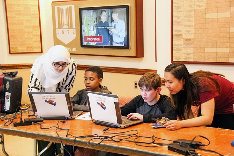 Two female adult volunteers helping two male children learn a new technology skill on laptop computers at Kid's Tech event