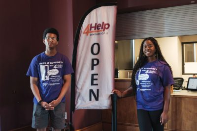 Two student workers wearing purple 4Help IT support shirts stand beside a tall “4Help Division of IT – OPEN” sign inside a service desk area.