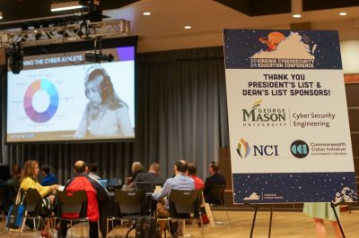 Attendees sit watching a presentation at the Virginia Cybersecurity Education Conference, with a sponsor sign in the foreground displaying logos for George Mason University, NCI, and the Commonwealth Cyber Initiative.