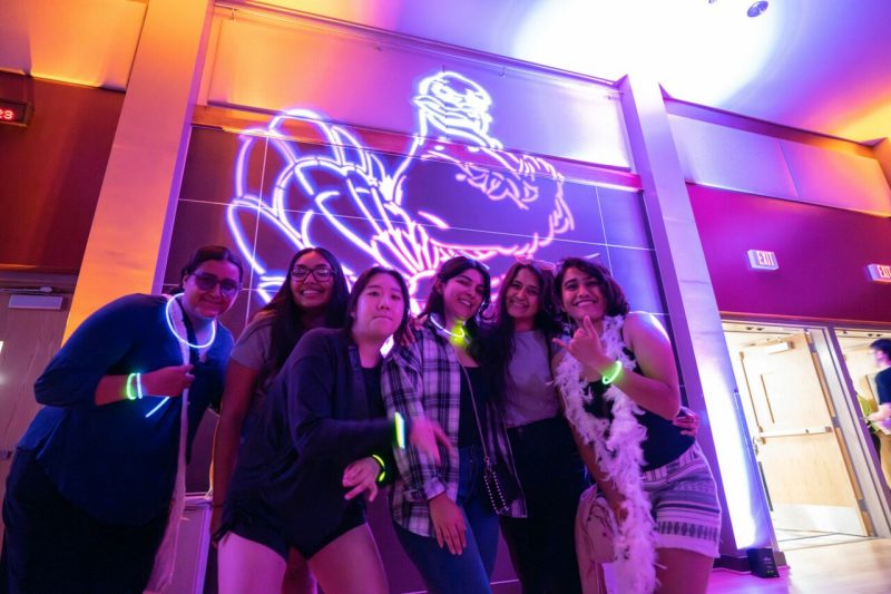 Students pose together at a glow-themed event with vibrant purple and orange lighting and a large illuminated HokieBird outline projected on the wall behind them.