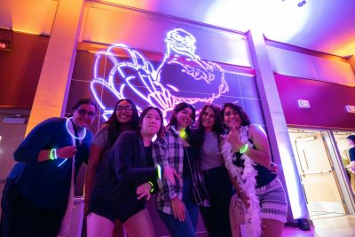 Students pose together at a glow-themed event with vibrant purple and orange lighting and a large illuminated HokieBird outline projected on the wall behind them.