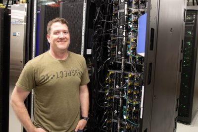 A man stands smiling in a server room next to racks of high-performance computing equipment and cooling systems.