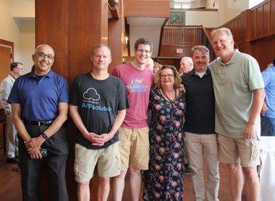 IT colleagues and friends standing together and smiling for a group photo at a May 2025 reception, gathered inside a wood-paneled event space.
