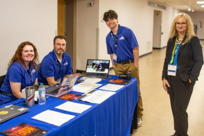 Four staff members stand and sit at an information table with laptops, printed materials, and a display screen, smiling for the camera at an event.
