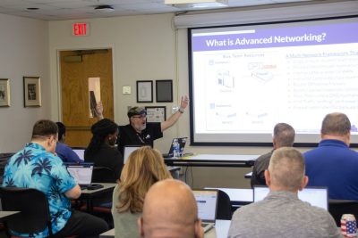Instructor gestures while teaching a classroom of adults during an advanced networking session, with a presentation slide displayed at the front of the room.