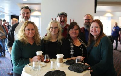Virginia Cyber Range colleagues gathered around a table at the 2024 Retiree Recognition & Holiday Social, smiling for a group photo as one person playfully makes bunny ears in the background.