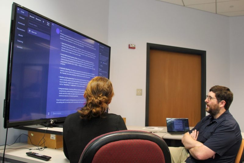 two people sit in front of a large digital screen in conversation.