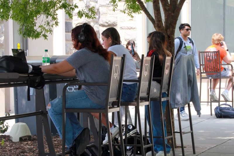 row of students sitting outdoors using laptops and phones