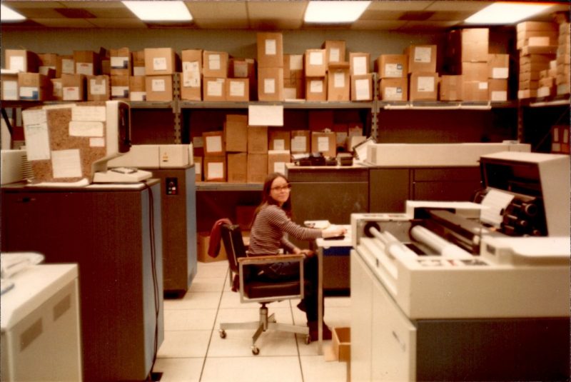 woman wearing glasses sits at a terminal in a computer room in the 1980s