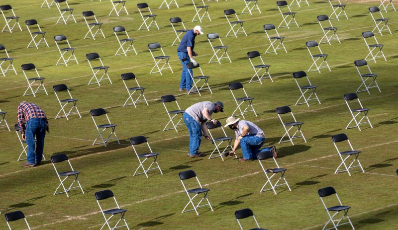 four people wearing blue jeans set up rows of evenly-spaced, physically-distanced chairs on grassy field