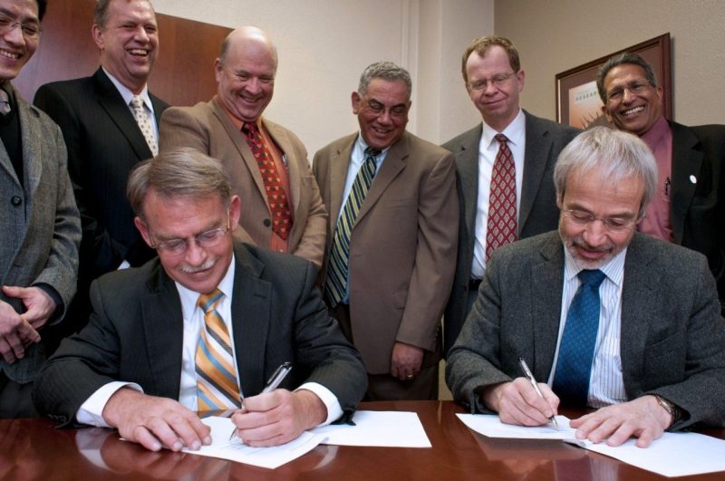 two people sign papers with onlookers smiling
