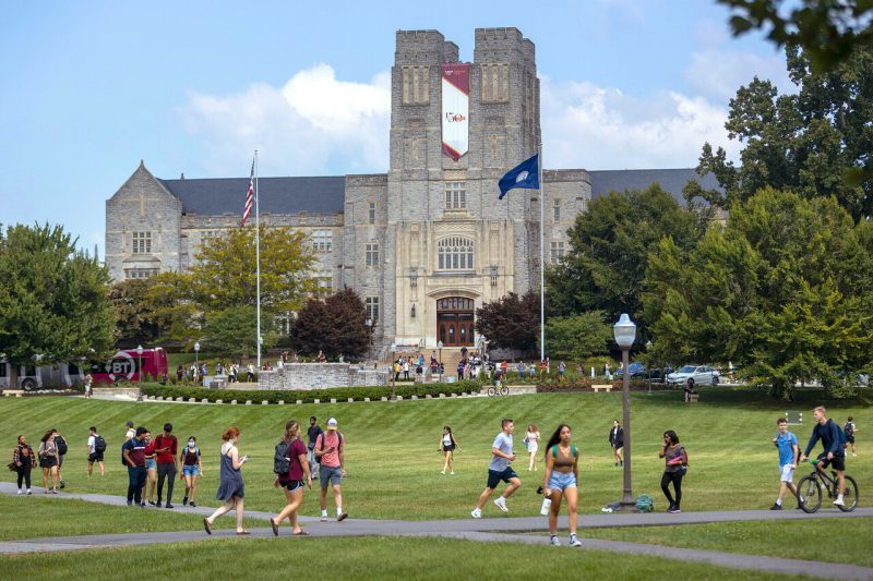 students walking across large field in front of stone building on clear summer day