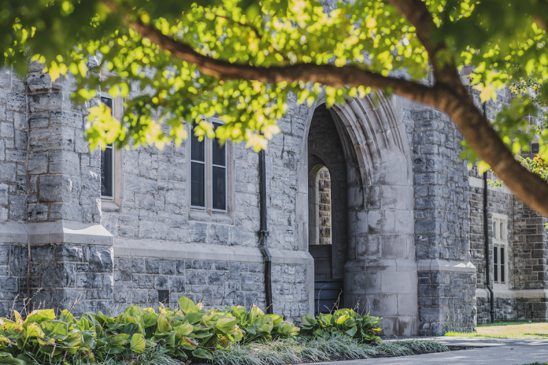 Grey Hokie Stone building featuring an archway. Tree limbs spread across the foreground. 